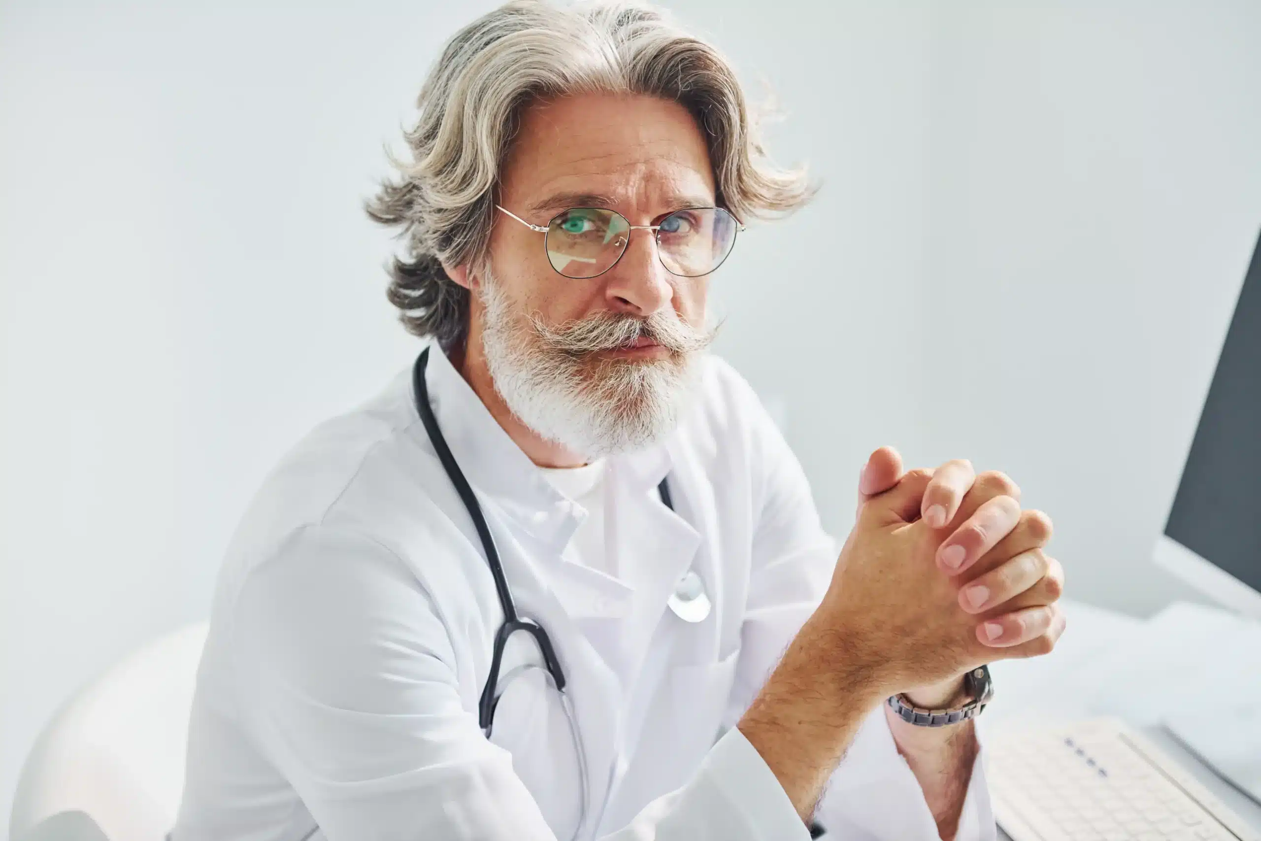 A imagem mostra um médico sênior, com expressão sérIa, em seu consultório. Ele olha para a câmera e veste jaleco e estetoscópio em volta do pescoço. Ilustra o texto sobre seguro erro médico.