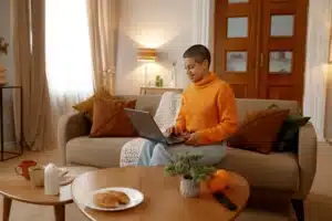 La imagen muestra a una joven sonriente sentada en un sofá en su casa. Tiene una computadora portátil en el regazo e ilustra el texto sobre la renovación de la Tarjeta Nacional de Registro Migratorio.