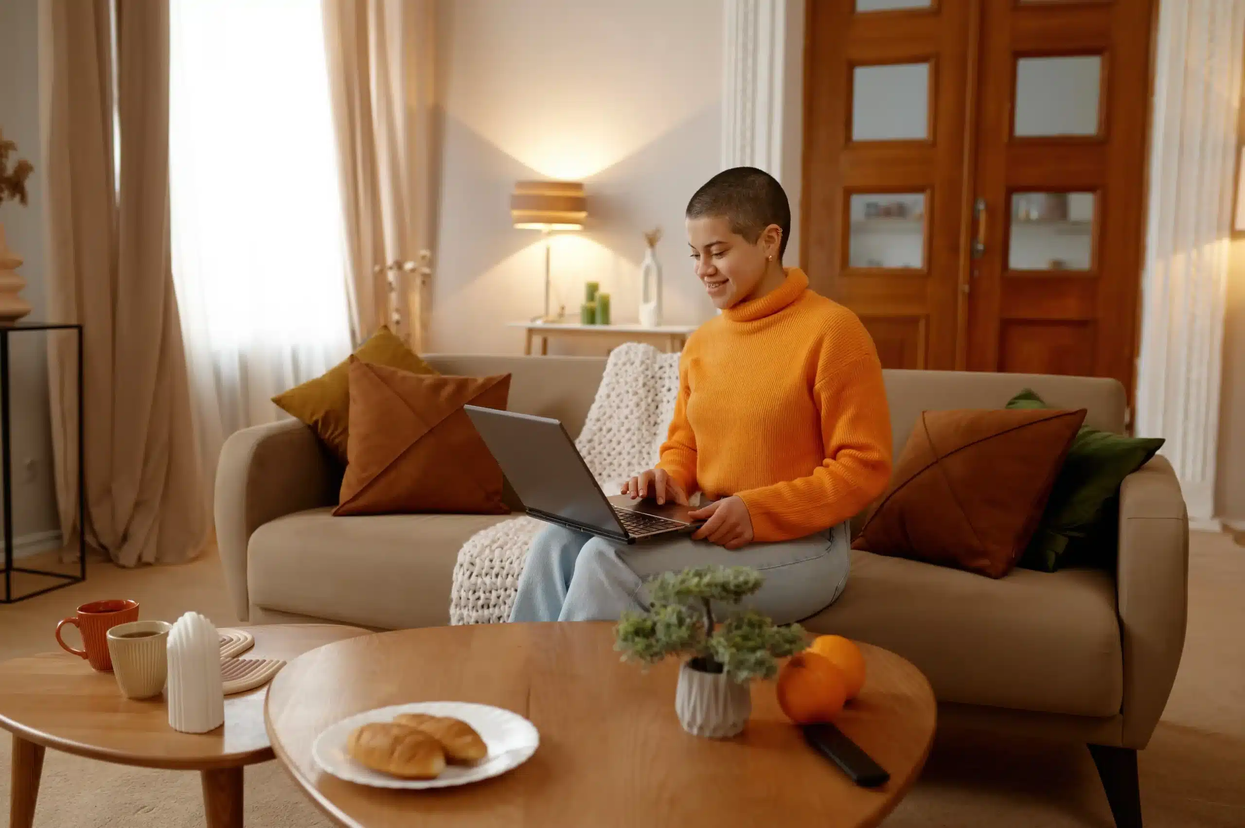 La imagen muestra a una joven sonriente sentada en un sofá en su casa. Tiene una computadora portátil en el regazo e ilustra el texto sobre la renovación de la Tarjeta Nacional de Registro Migratorio.
