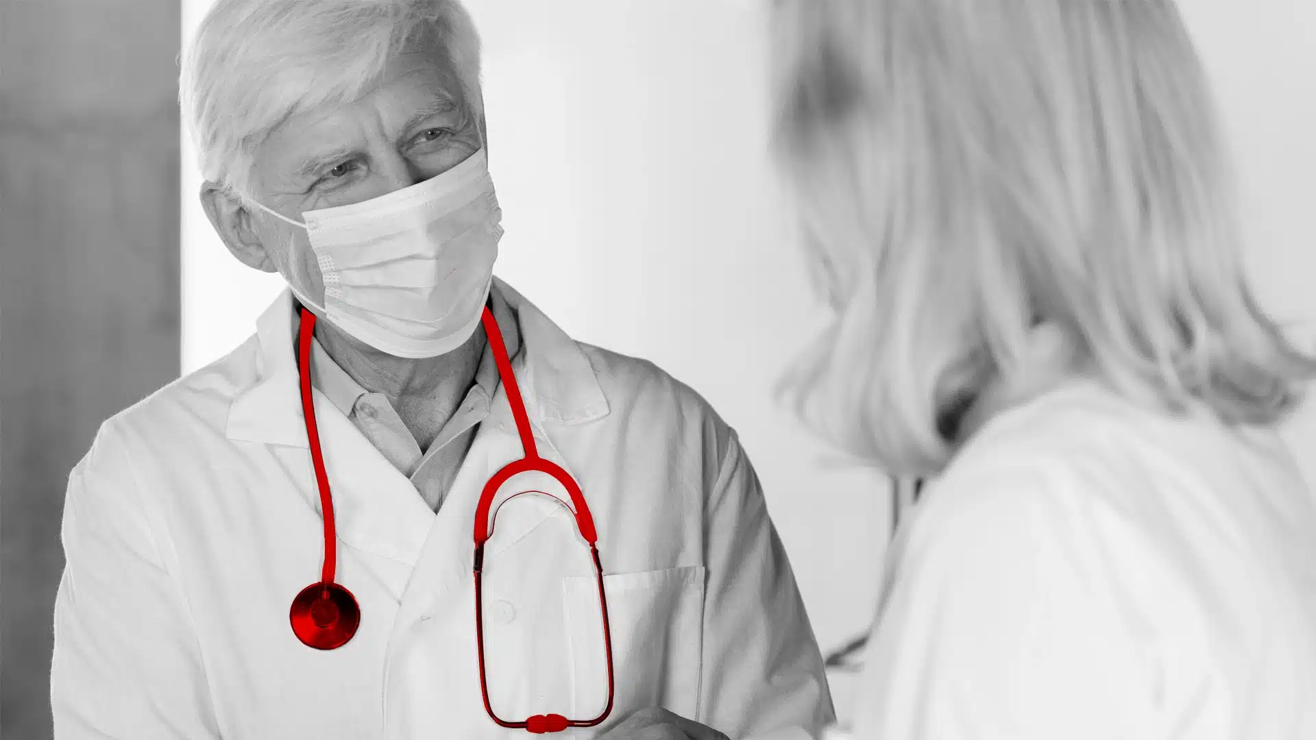 A imagem mostra um médico idoso, utilizando jaleco, estetoscópio e máscara, enquanto conversa com uma mulher.