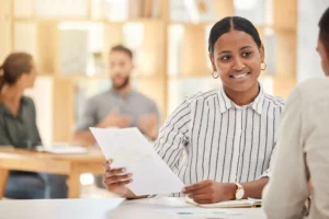 La imagen muestra a una joven en un entorno corporativo, con documentos en la mano y sonriendo. Ilustra el texto sobre la contratación de refugiados.