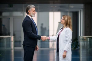 A imagem mostra um advogado, de meia idade, usando terno e sorrindo, apertando as mãos de uma médica, de jaleco. Ambos estão fechando um negócio e ilustra o texto contratar advogado em Direito Médico.
