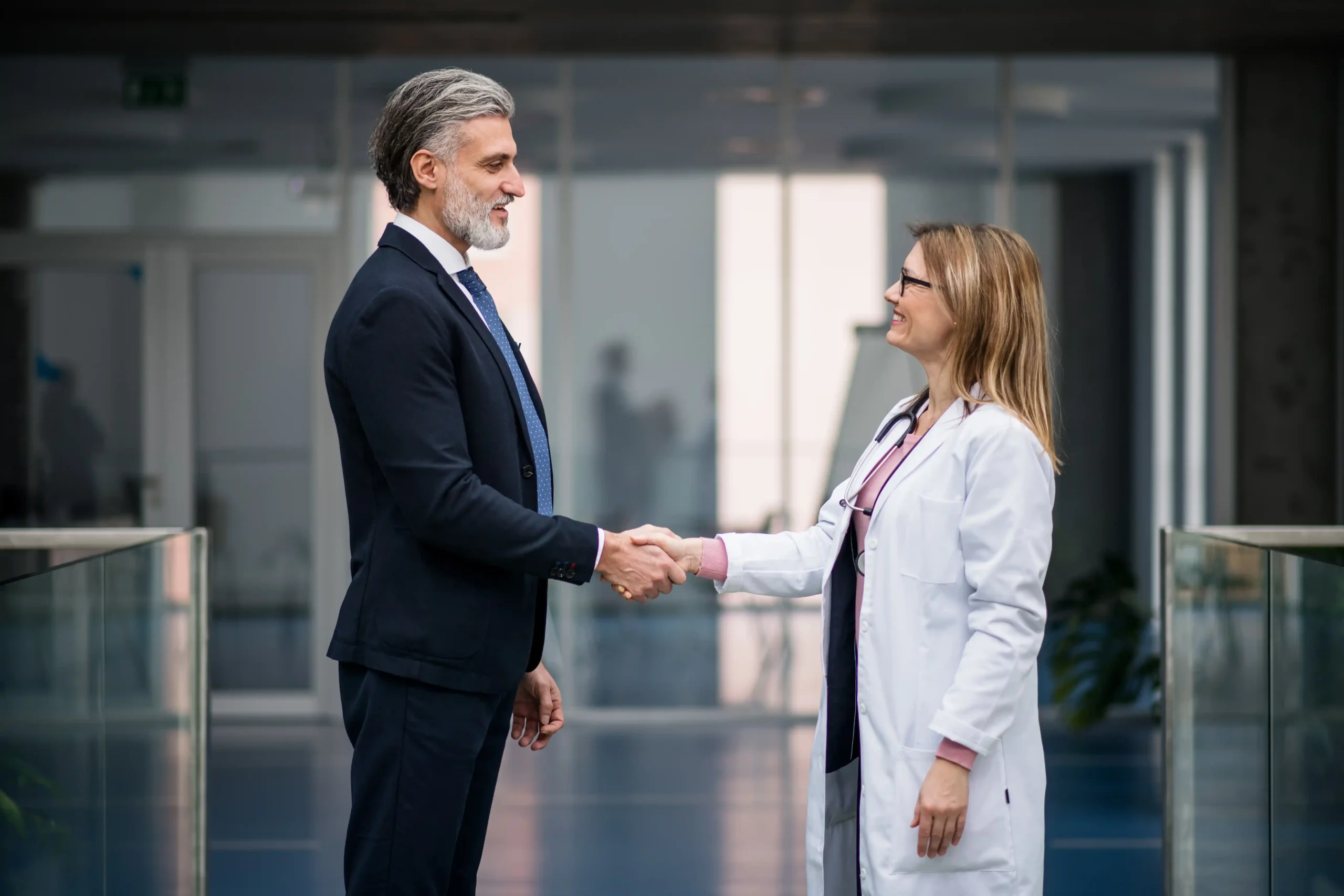 A imagem mostra um advogado, de meia idade, usando terno e sorrindo, apertando as mãos de uma médica, de jaleco. Ambos estão fechando um negócio e ilustra o texto contratar advogado em Direito Médico.