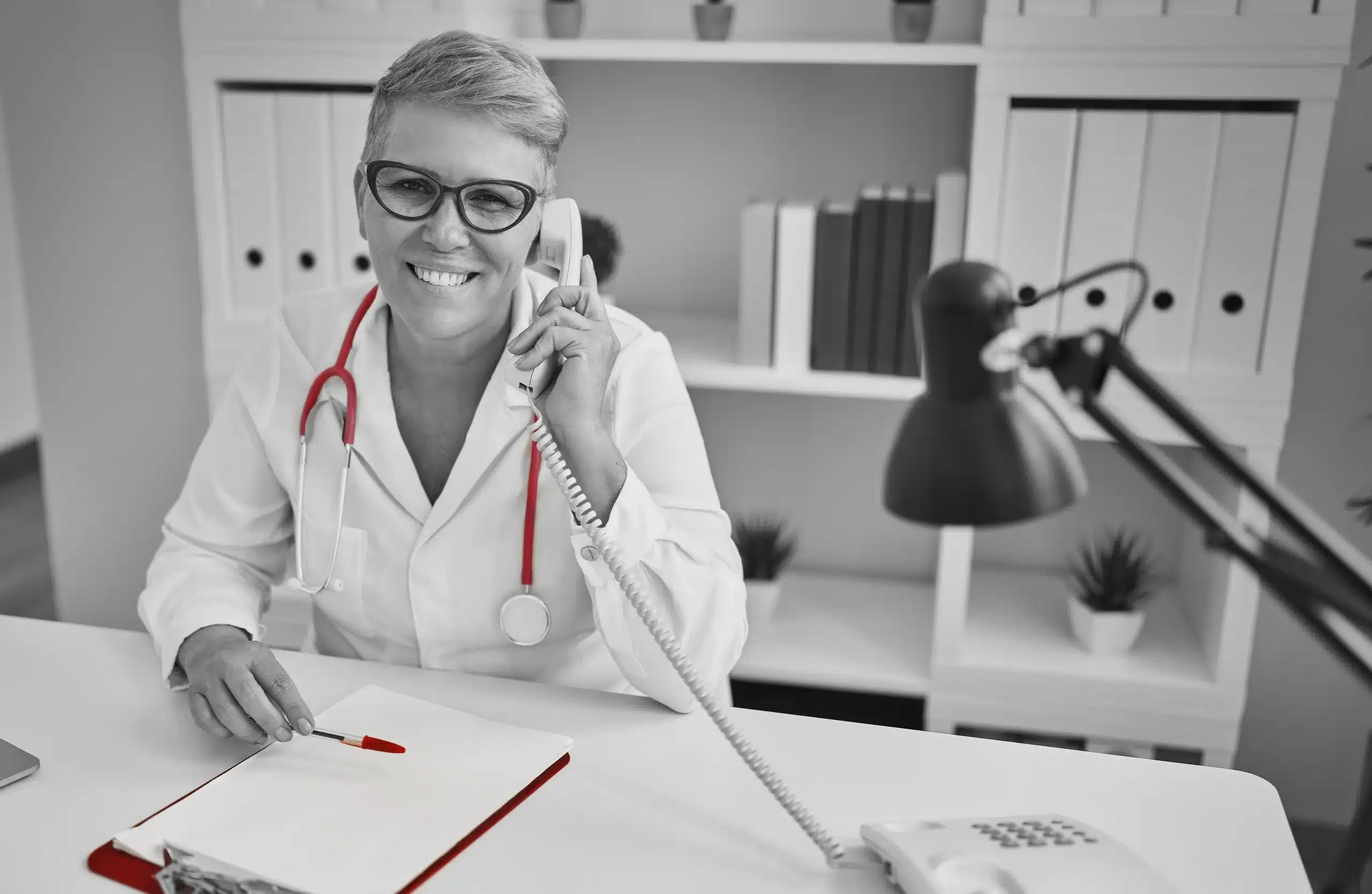 A imagem mostra uma médica, sorrindo para a câmera, enquanto utiliza o telefone. Ela está sem seu consultório, sentada em uma mesa.