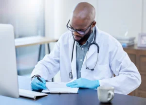 A imagem mostra um médico, de jaleco, usando estetoscópio em volta do pescoço, sem seu consultório. Ele lê documentos e possui uma caneta em uma das mãos. Ilustra o texto sobre modelo de defesa de sindicância CRM.