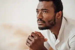 La imagen muestra a un joven negro, pensativo, mirando al horizonte con las manos entrelazadas. Ilustra el texto sobre los refugiados en Brasil.