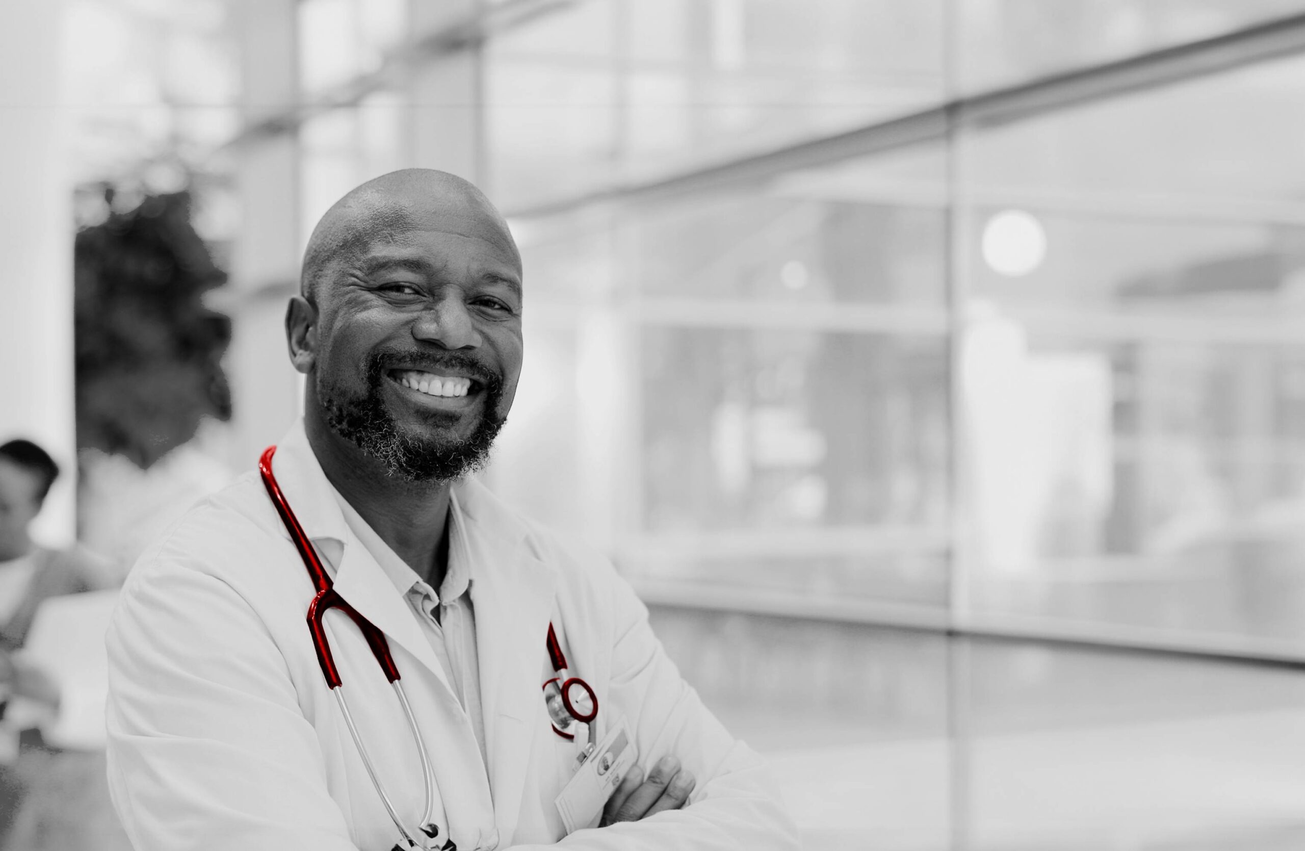 A imagem mostra um jovem médico sorrindo para a câmera, ele utiliza jaleco e estetoscópio e e está em ambiente hospitalar.