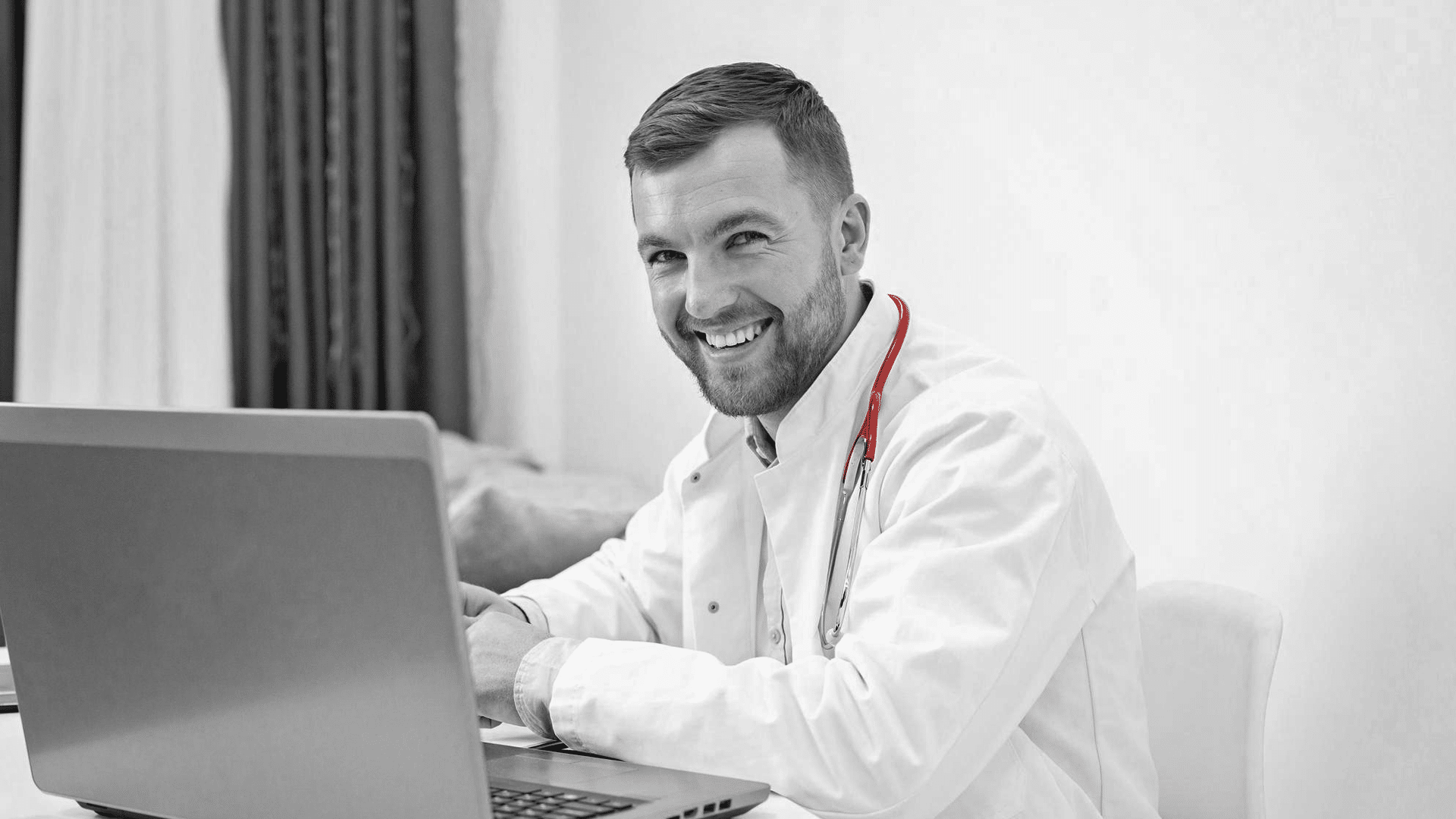 A imagem mostra um médico jovem, de jaleco e estetoscópio, sorrindo para câmera, utilizando o laptop.