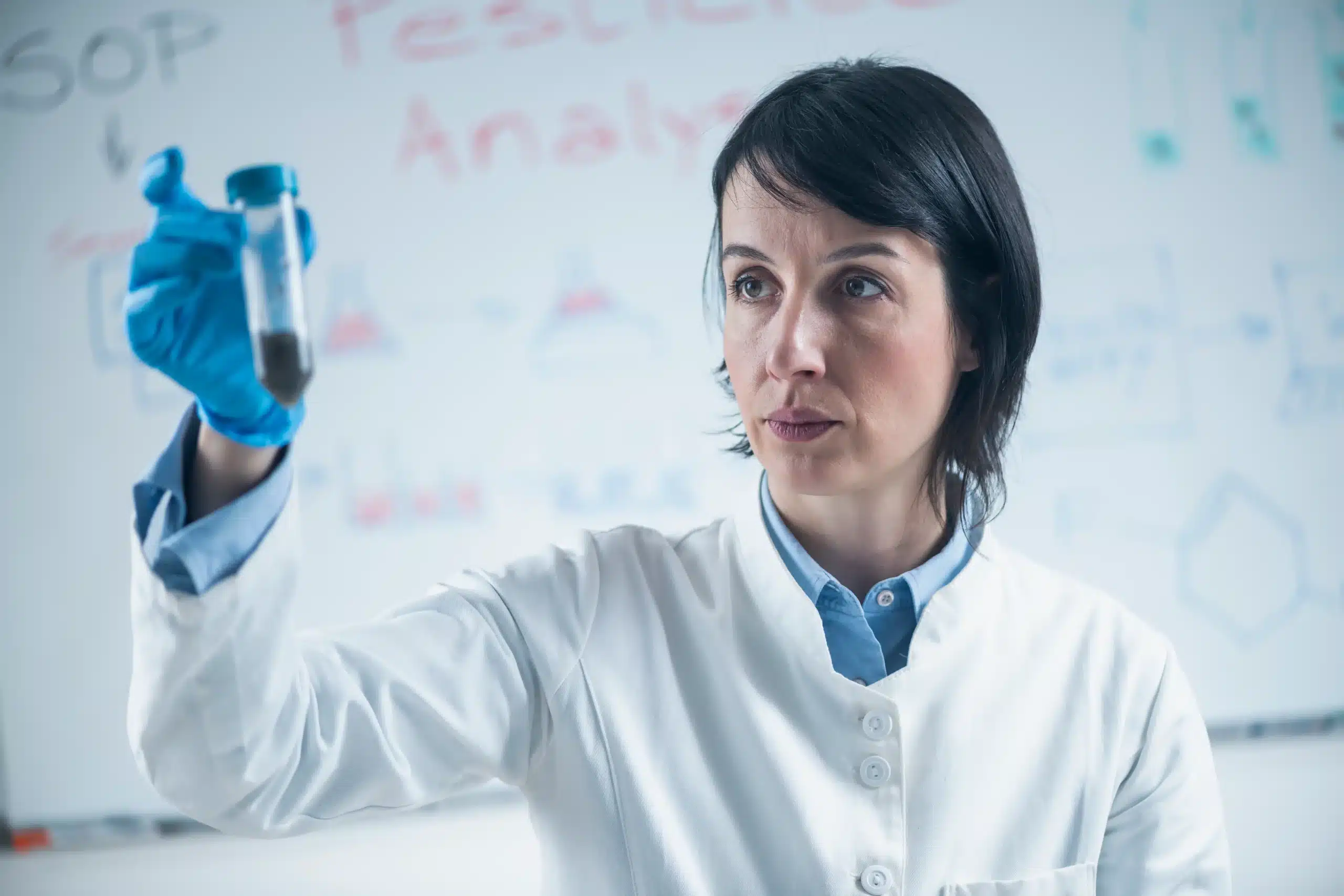 La imagen muestra a un científico, con bata de laboratorio, en un laboratorio, analizando un tubo de ensayo. Ilustra el texto sobre visa ​​para investigación científica.