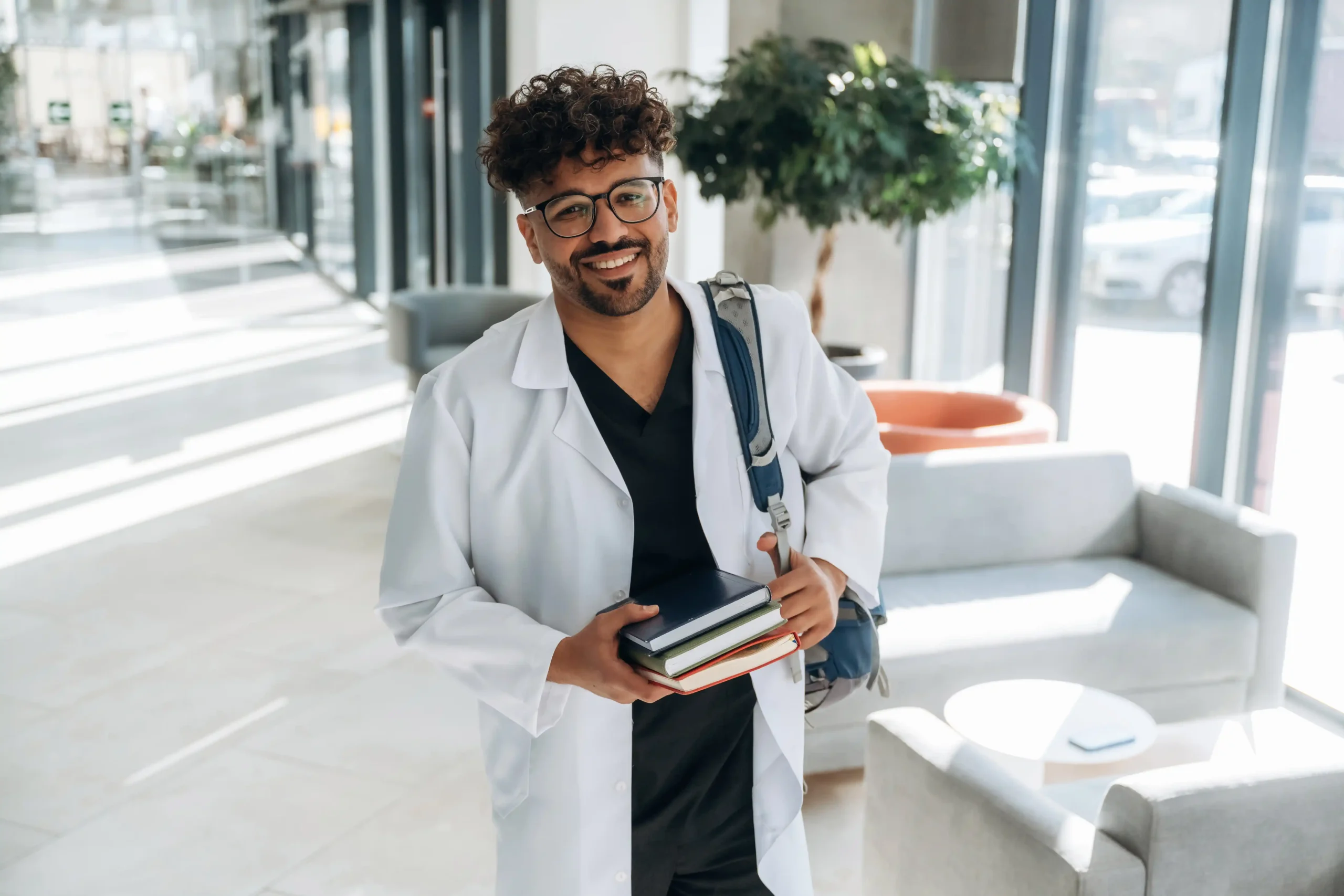 The image shows a young doctor, wearing a lab coat, smiling at the camera while carrying books in one hand. It illustrates the text about The Visa for Medical Training.