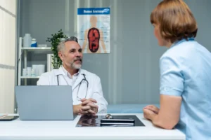 A imagem mostra um médico de meia-idade, em seu consultório, atendendo uma paciente. Ilustra o texto sobre contrato entre médico e paciente.