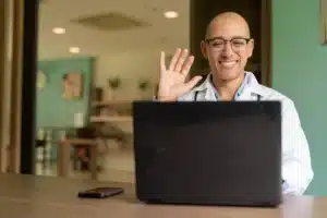 A imagem mostra um médico, de jaleco, estetoscópio, em frente ao laptop, sorrindo e acenando, conversando com um paciente. Ilustra o texto sobre telemedicina.