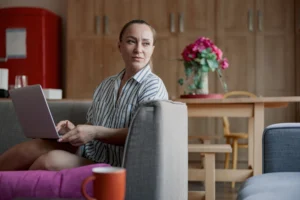 La imagen muestra a una mujer, con expresión seria, sentada en su sofá con su portátil en el regazo. Ilustra el texto sobre visa de turista para los estadounidenses.