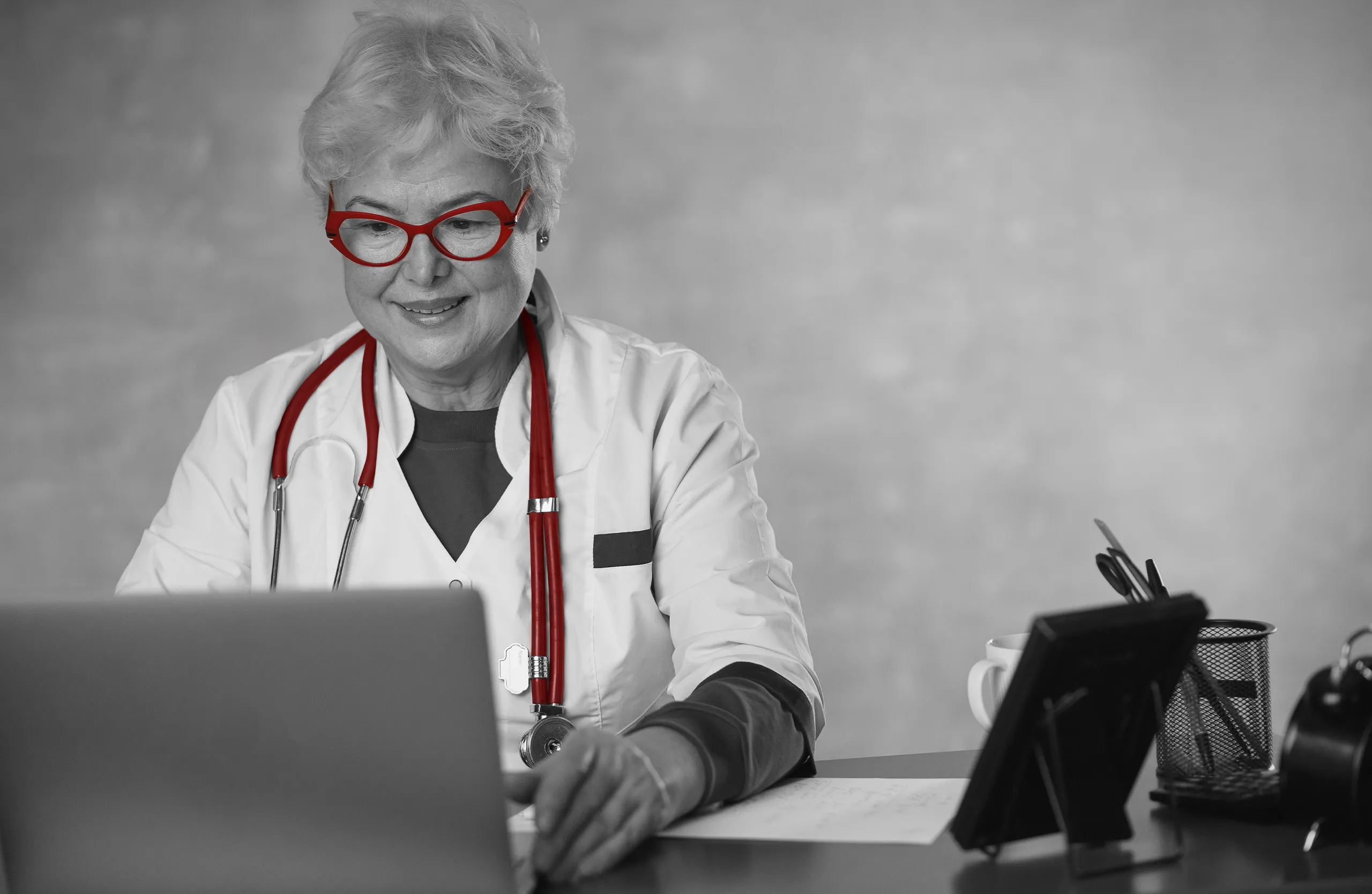 A imagem mostra uma médica sênior, sorrindo, enquanto utiliza o laptop. Ela usa jaleco e estetoscópio e está em seu consultório.