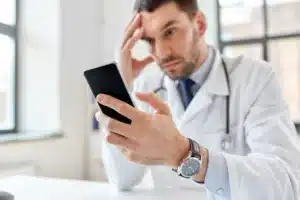 A imagem mostra um jovem médico, segurando um smartphone, com expressão preocupada, com a outra mão segurando o rosto. Ilustra o texto sobre crise de imagem profissional do médico.