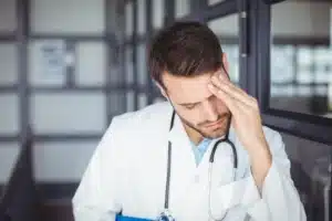 A imagem mostra um jovem médico de jaleco, estetoscópio, preocupado, com uma das mãos na testa. Ilustra o texto sobre judicialização da saúde.