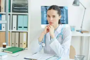 A imagem mostra uma jovem médica usando jaleco, estetoscópio, em seu consultório, com expressão séria, olhando para a câmera. Ilustra o texto sobre iIatrogenia,