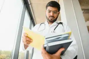 A imagem mostra um jovem médico, usando jaleco e estetoscópio, analisando documentos e cadernos. Ilustra o texto sobre médico imposto de renda.