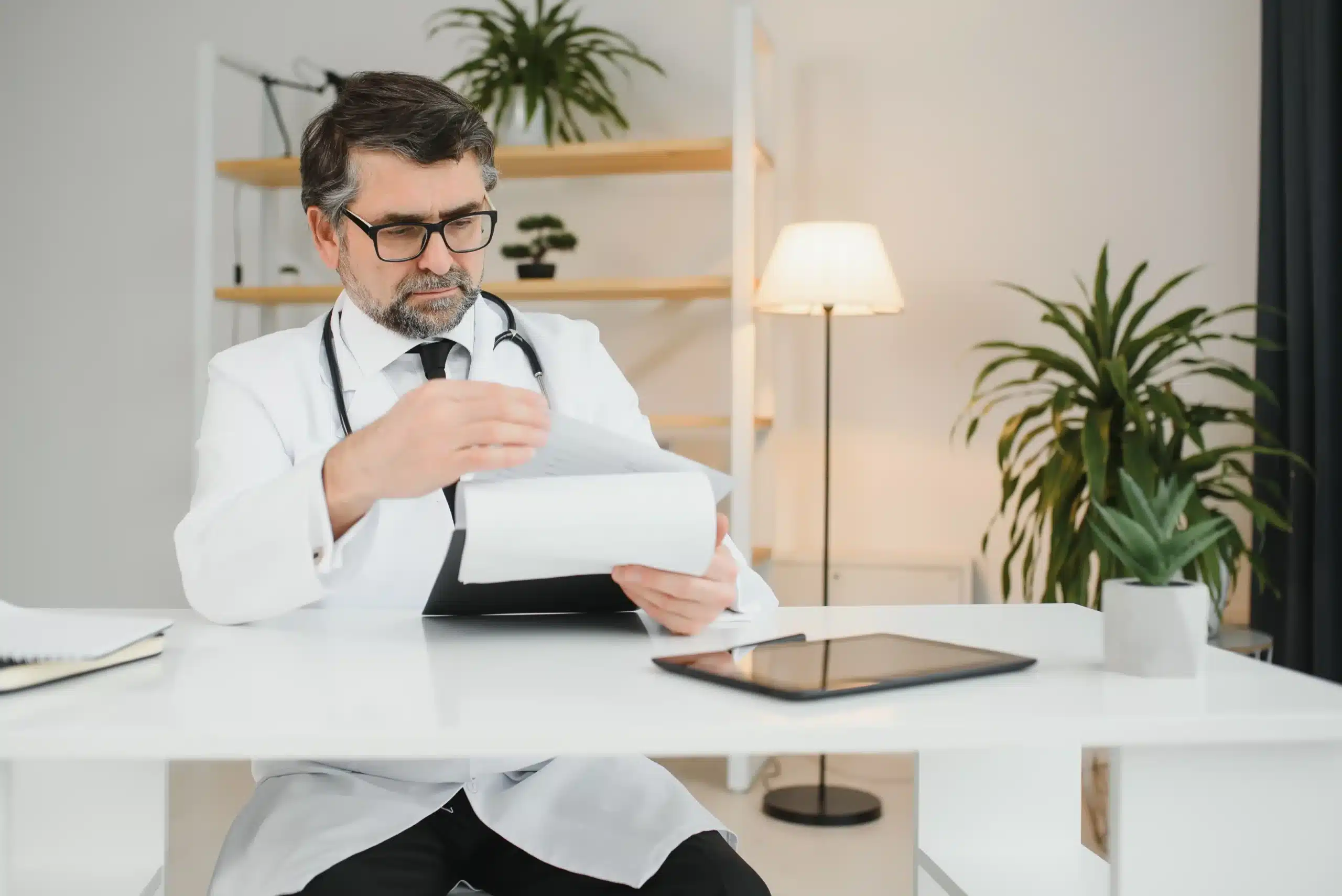 A imagem mostra um médico de meia-idade, com jaleco, gravata e estetoscópio em volta do pescoço, em seu consultório. Ele analisa documentos. Ilustra o texto sobre médico e imposto.