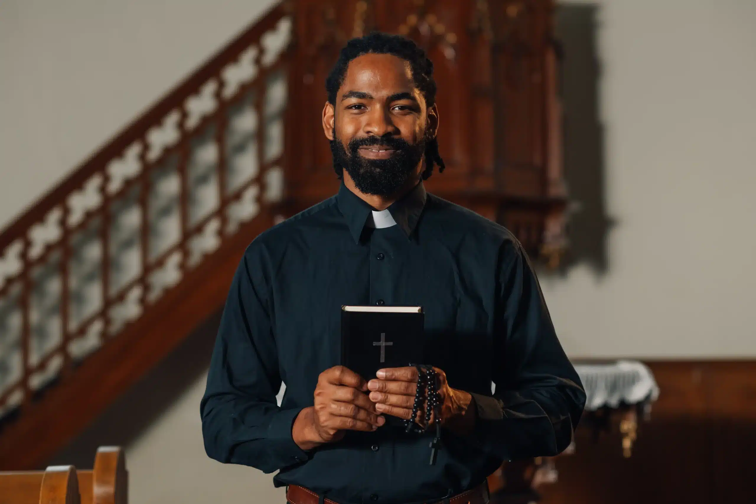 La imagen muestra a un joven sacerdote sosteniendo una Biblia y un rosario en la iglesia, mirando a la cámara y sonriendo. Ilustra el texto sobre la visa para actividades religiosas en Brasil.