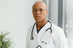 A imagem mostra um médico jovem, usando jaleco, estetoscópio em volta do pescoço, com expressão séria, olhando para a câmera. Ilustra o texto sobre erro médico ou iatrogenia.