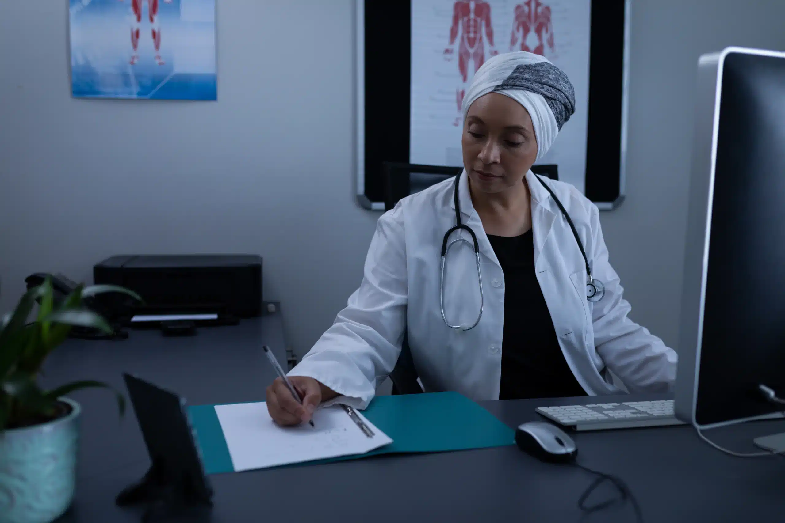 Médica usando jaleco e estetoscópio, sentada à mesa de consultório, escrevendo em uma folha enquanto olha para o documento. Ao fundo, há pôsteres anatômicos e um computador iluminado ao lado dela.