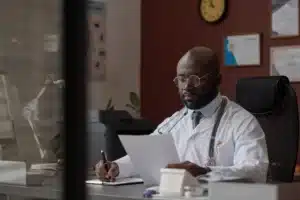 Médico negro usando óculos e jaleco branco trabalha em seu consultório, analisando documentos sobre a mesa. Ele está sentado em uma cadeira de escritório, com um estetoscópio no pescoço, concentrado na leitura e escrita de papéis. Ao fundo, há certificados na parede, um relógio e equipamentos de escritório como impressora e luminária.