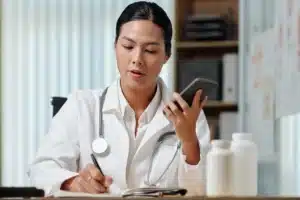 A imagem mostra uma jovem médica em seu consultório, usando jaleco e estetoscópio em volta do pescoço. Ela faz anotações com papel e caneta e, na outra mão, segura um smartphone. Ilustra o texto sobre imposto para médicos.