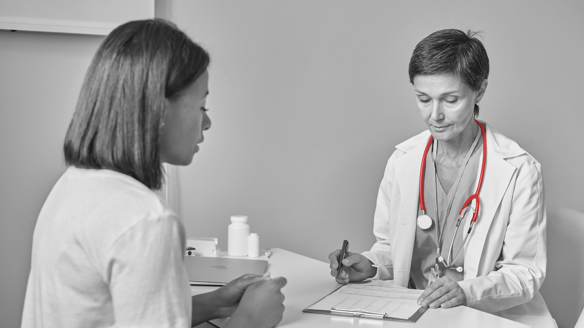 A imagem mostra uma médica de meia-idade, usando jaleco e estetoscópio em volta do pescoço, atendendo uma paciente em consultório.