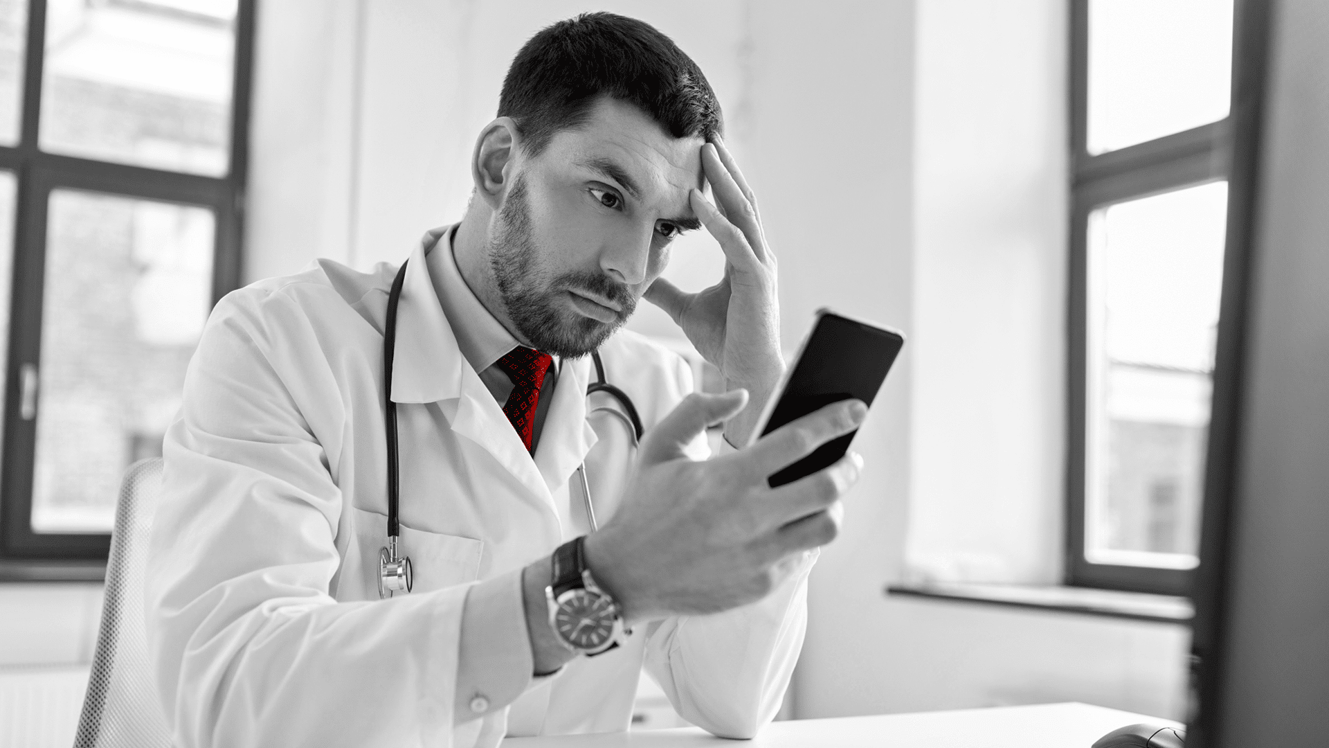 A imagem mostra um jovem médico, de jaleco, estetoscópio em volta do pescoço, preocupado, segurando com uma das mãos um smartphone e outra, a testa.