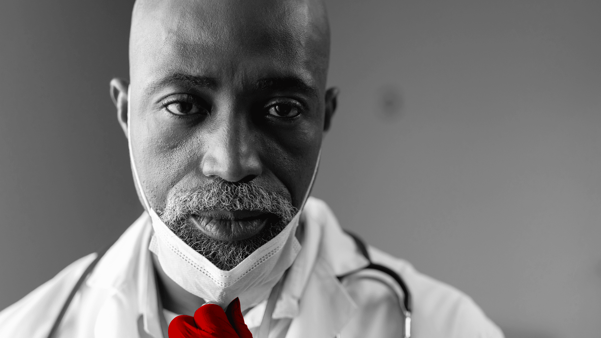 A imagem mostra em destaque o rosto de um médico de meia-idade, de lucas cirúrgicas, jaleco e estetoscópio em volta do pescoço. Ele puxa a máscara com uma das mãos, com expressão séria.