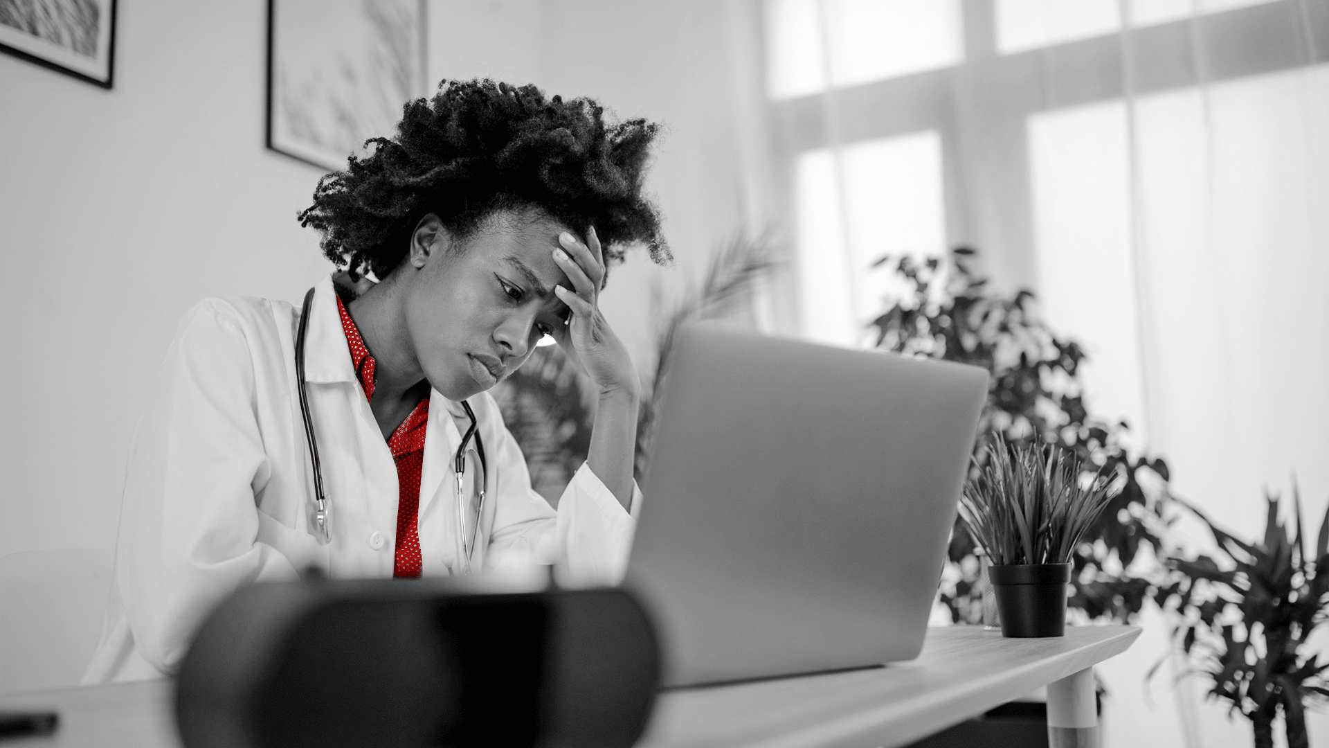 A imagem mostra uma jovem médica, preocupada, com uma das mãos em sua testa. Ela está em seu consultório, usando jaleco e estetoscópio em volta do pescoço.