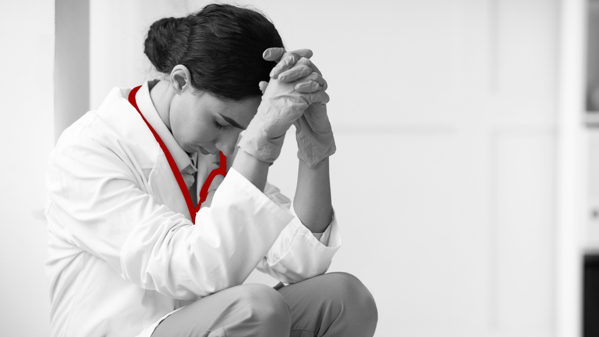 A imagem mostra uma jovem médica, de jaleco, estetoscópio em volta do pescoço. Ela está preocupada, sentada contra a parede.