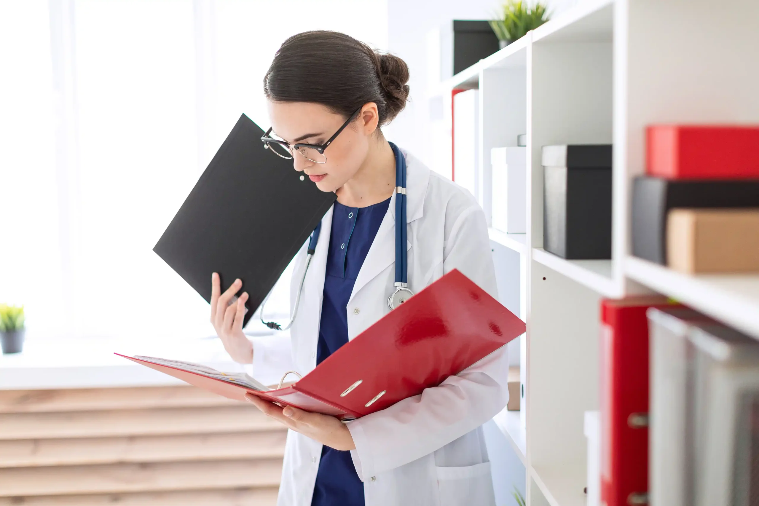 A imagem mostra uma jovem médica, usando jaleco, com estetoscópio em volta do pescoço, checando documentos em seu consultório. Ilustra o texto sobre imposto para médicos e profissionais da saúde.