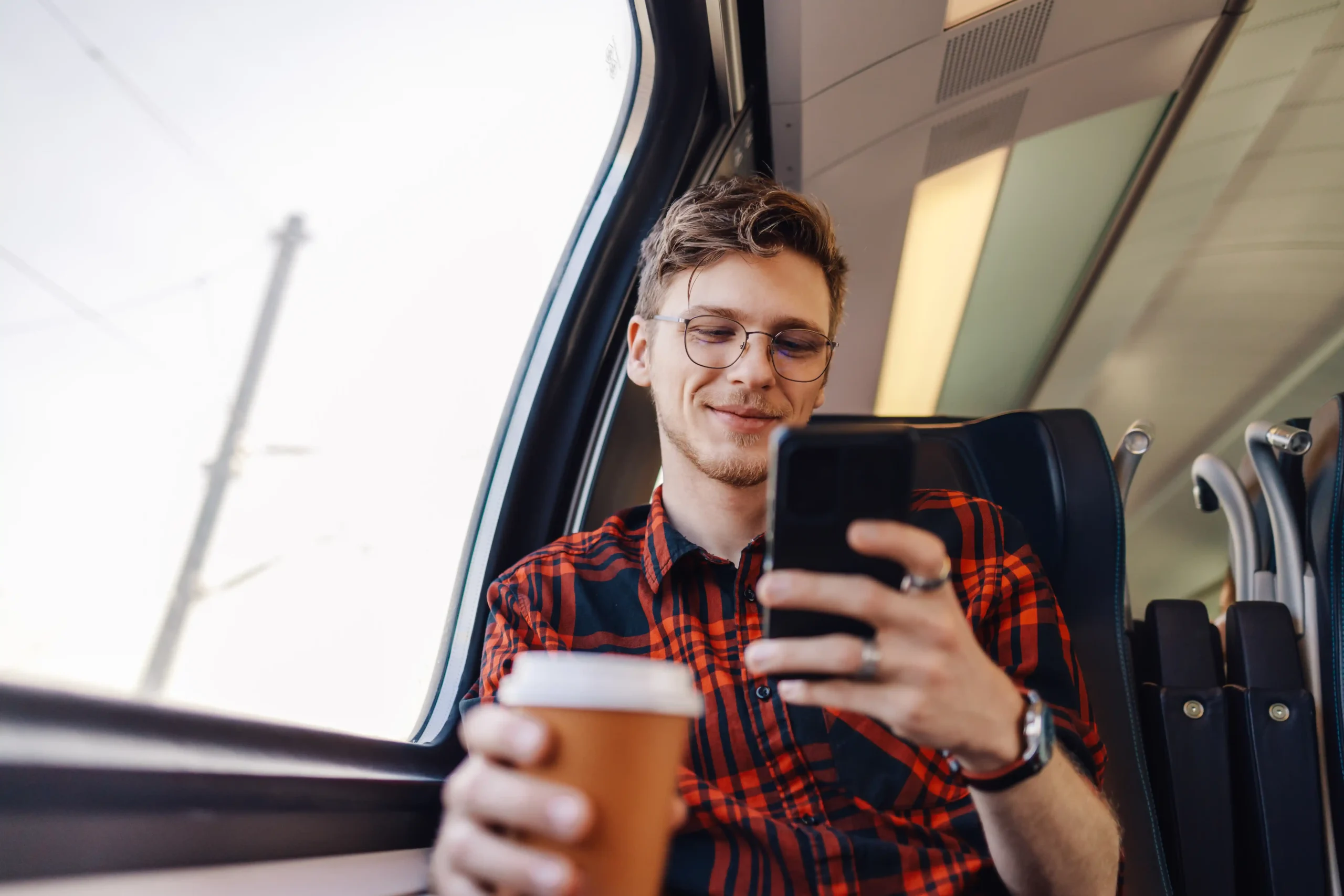 La imagen muestra a un joven viajando en tren, sentado y sonriendo con su teléfono inteligente. Ilustra el texto sobre el Programa de Trabajo y Vacaciones en Brasil.