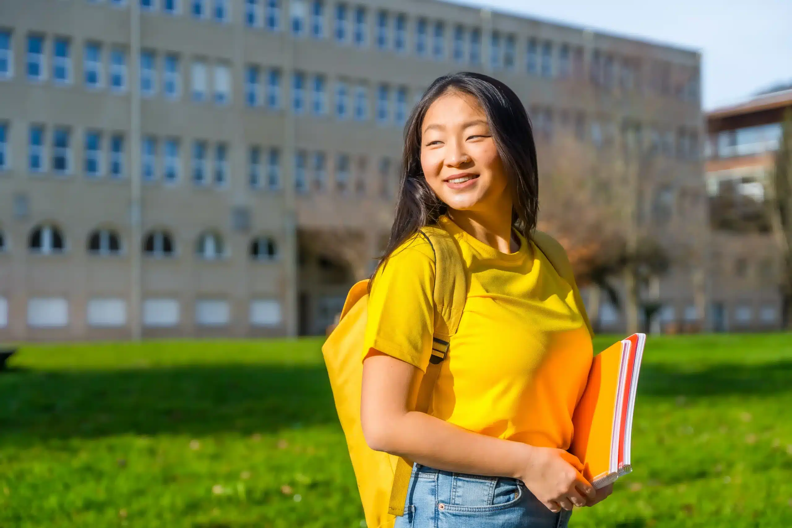 La imagen muestra a una joven estudiante sonriendo y sosteniendo cuadernos. Está al aire libre en su campus. Ilustra el texto sobre visas para prácticas académicas en Brasil.