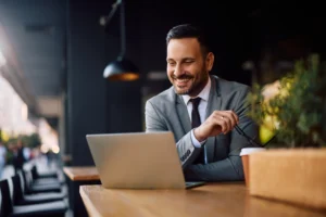 The image shows a smiling young entrepreneur using a laptop in a café. The image illustrates the text about the Brazilian Visa for Activities of Relevance.