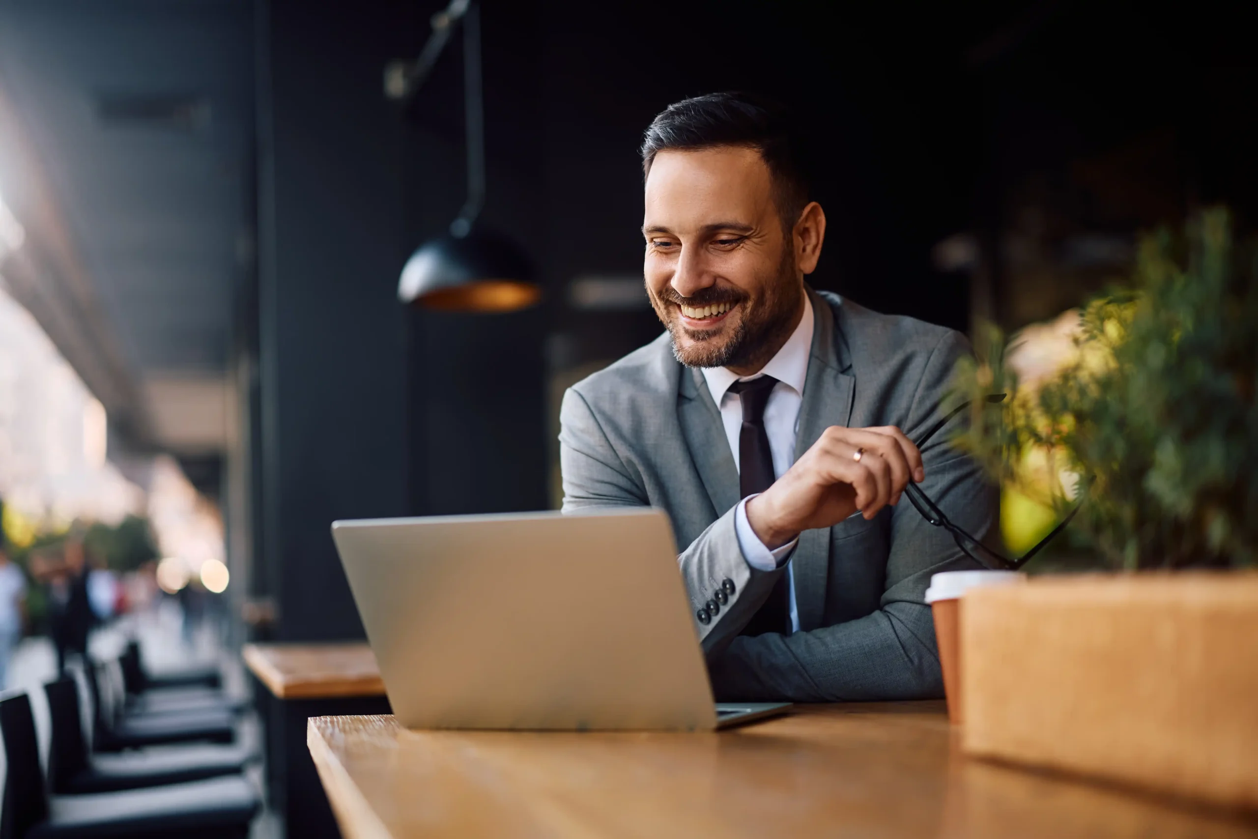 The image shows a smiling young entrepreneur using a laptop in a café. The image illustrates the text about the Brazilian Visa for Activities of Relevance.