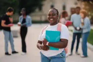 The image shows a young student smiling at the camera while holding a notebook. She is outdoors on her campus. It illustrates the text about visas for academic internships in Brazil.