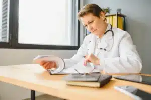 A imagem mostra uma média de meia-idade, em seu consultório, analisando documentos. Ela usa jaleco e estetoscópio em volta do pescoço. Ilustra o texto sobre modelo de contrato clínica médico.
