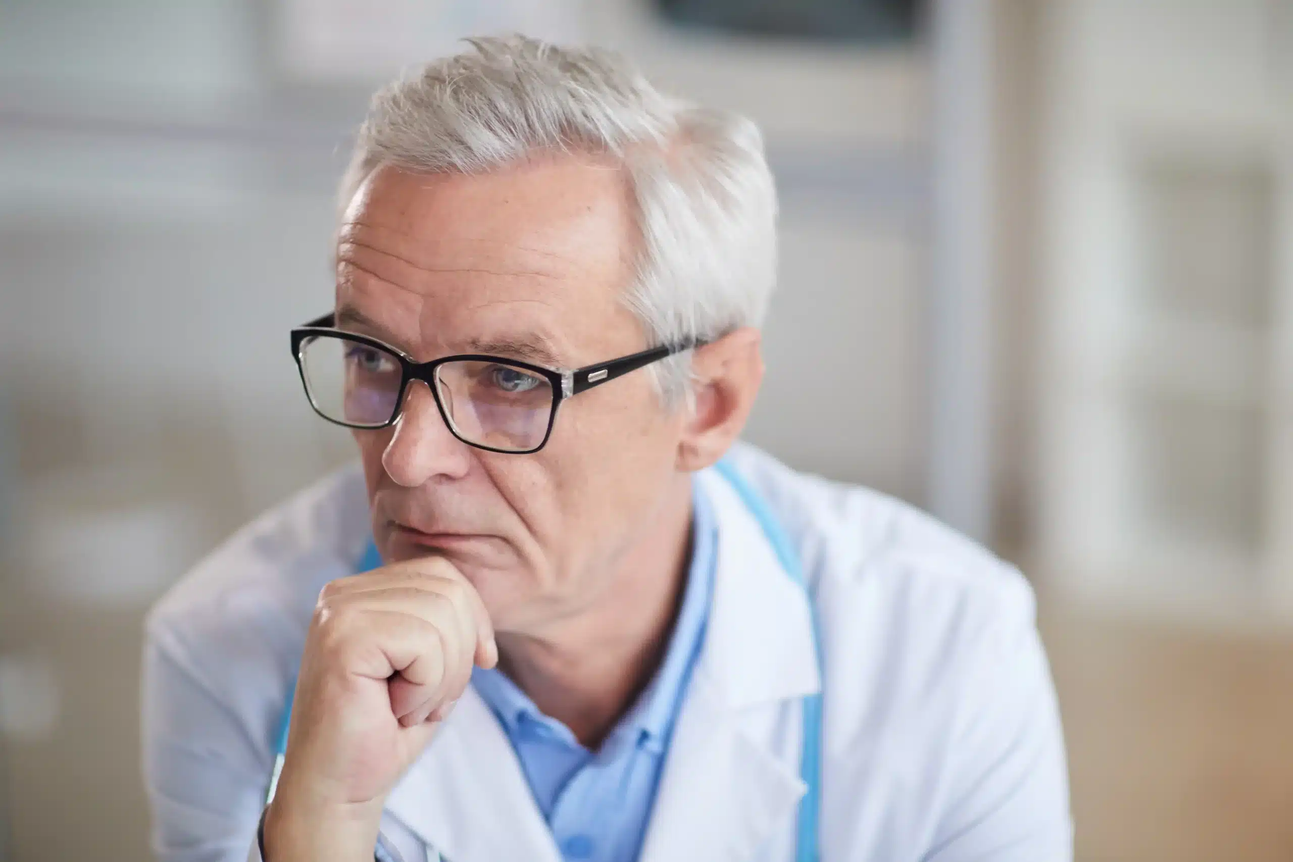 A imagem mostra um médico idoso, usando jaleco, estetoscópio em volta do pescoço. Ele apoia o queixo com uma das mãos, com expressão séria. Ilustra o texto sobre Defesa de sindicância no CRM.