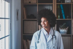 A imagem mostra uma jovem médica, sorridente, em seu consultório. Ela usa jaleco e estetoscópio em volta do pescoço.