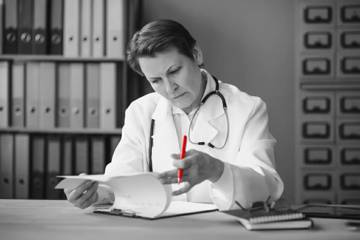 A imagem mostra uma médica, em seu consultório, sentada à mesa, verificando documentos.