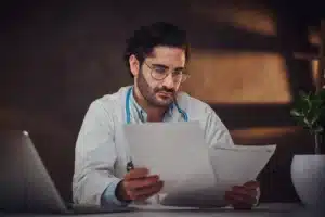 A imagem mostra um médico, de jaleco, estetoscópio em volta do pescoço, em se consultório, analisando papéis. Ilustra o texto sobre Imposto médico Reforma tributária.