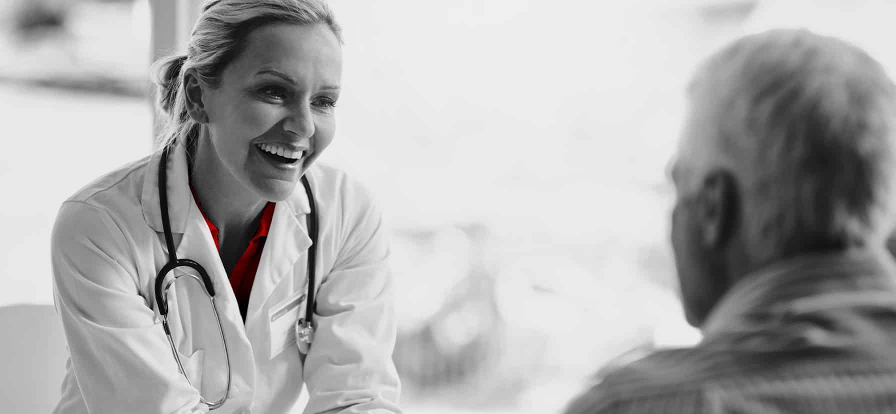 A imagem mostra uma médica, sorridente, de jaleco e estetoscópio em volta do pescoço, atendento um paciente e sorrindo.