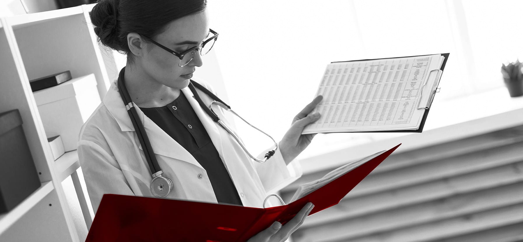 A imagem motra uma jovem médica, de jaleco, estetoscópio em volta do pescoço, em seu consultório, analisando documentos.