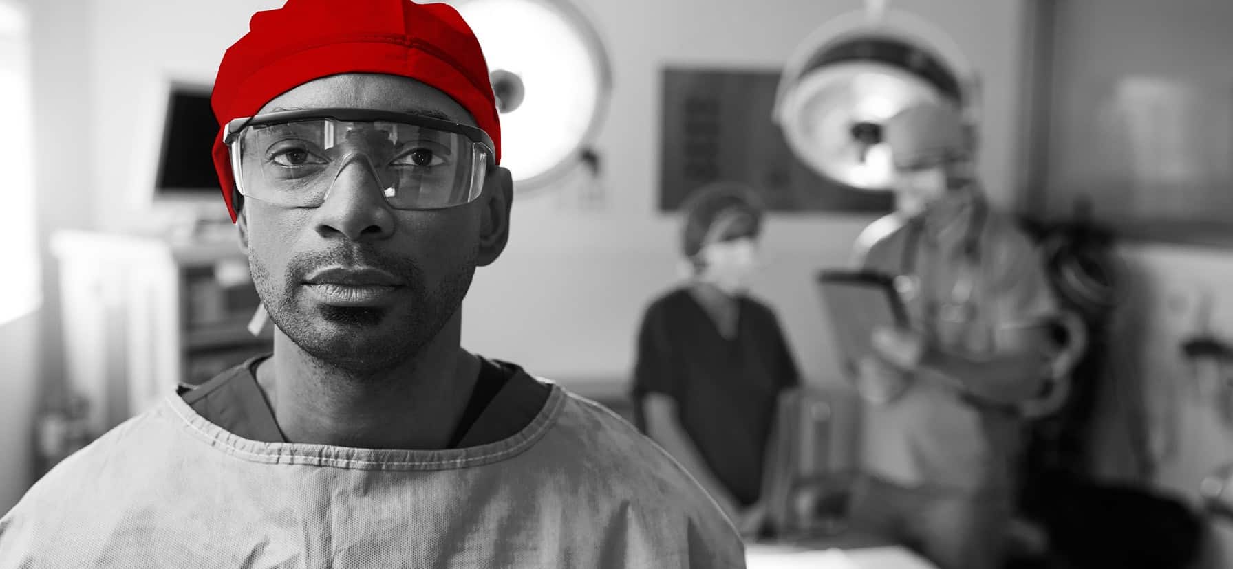 A imagem mostra um jovem médico, com roupas cirúrgicas, na sala de cirurgia, olhando com expressão séria para a câmera.