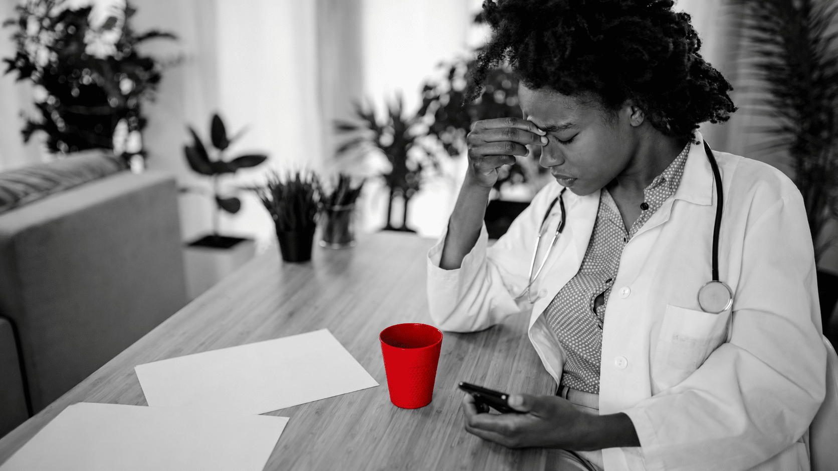 A imagem mostra uma jovem médica, usando jelaco e estetoscópio em volta do pescoço, sentada à mesa em seu consultório. Ela tem expressão de preocupação, segurando a testa com uma das mãos.