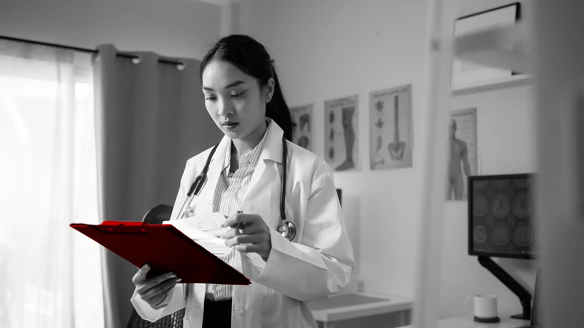 A imagem mostra uma médica, em seu consultório, usando jaleco e estetoscópio em volta do pescoço, verificando uma prancheta.