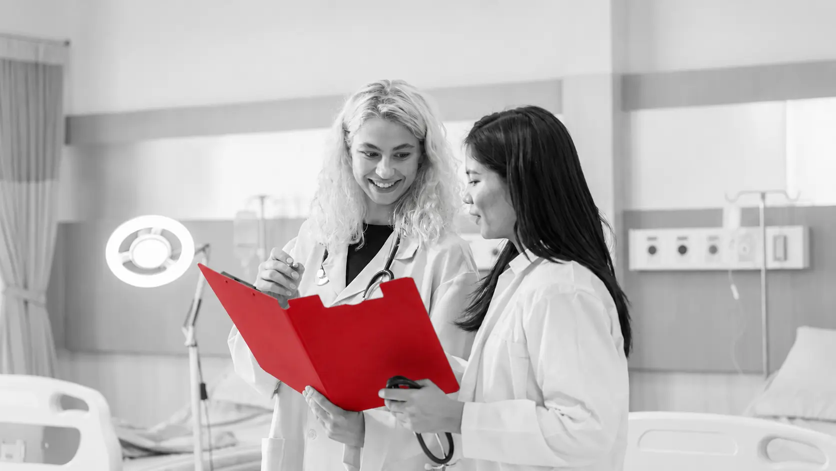 A imagem mostra duas médicas, usando jaleco e estetoscópio em volta dos pescoços. Ambas estão analisando um prontuário médico em ambiente hospitalar.